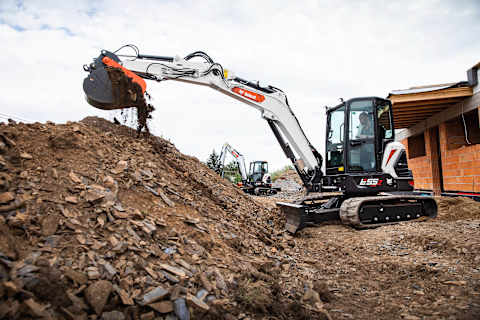 Two male operators using Bobcat excavators, E55z and E50Z, to scoop debris on an overcast day.