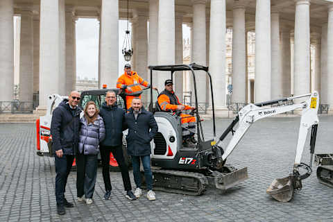 A group of people smiling in front of two Bobcat excavators and operators, with the pillars of a coliseum in the background.