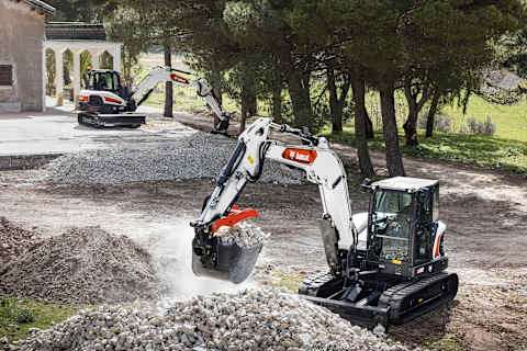 Two male operators using Bobcat excavators to move piles of stone in front of a house, with trees nearby.