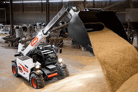 An action shot of a male operator using a Bobcat TL25.60 telehandler to unload grain into a cow barn.