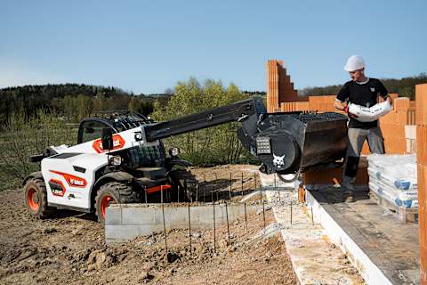 Imagen de un manipulador telescópico Bobcat TL25.60 con un trabajador cargando sacos de materiales desde una zona de almacenamiento.