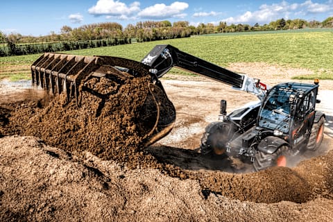 A smiling male operator uses a Bobcat TL38.70 telehandler to lift a massive pile of dirt, with a grassy field in the background.