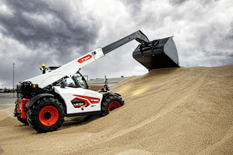 Снимок с видом сбоку на телескопический погрузчик Bobcat TL38.70HF, перемещающий большую кучу зерна на открытом воздухе в пасмурный день.