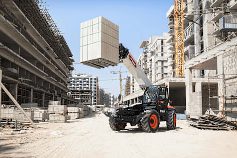 An action shot of a Bobcat TL40.180SLP telehandler lifting cinder blocks with its boom extended on a building construction site.