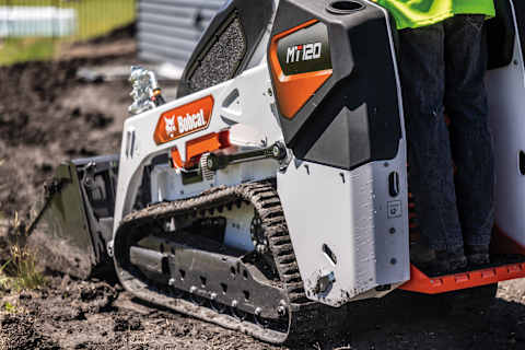 An operator stands on the platform of the Bobcat MT120 mini track loader that is working on a muddy job site.
