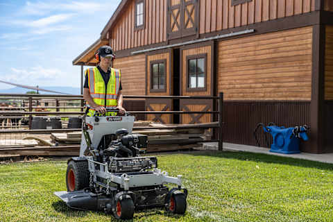 Professional operator mowing farm property with Bobcat ZS4000 stand-on zero-turn mower with brown wooden barn in the background