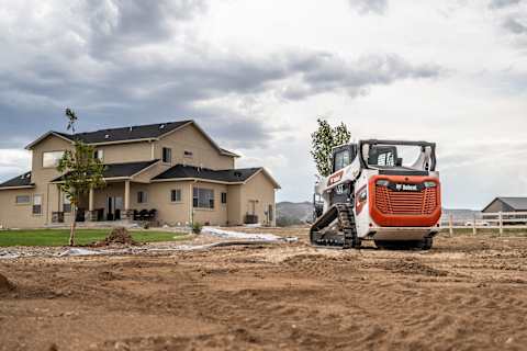 A Bobcat Compact Track Loader Travels on a Large Patch of Dirt on a Residential Development. A Large Home With Finish Landscaping Is in the Background.