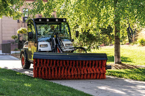 Front view of an operator sweeping the sidewalk at a college campus with an angle broom attached to a Bobcat UW53 Toolcat Utility Work Machine