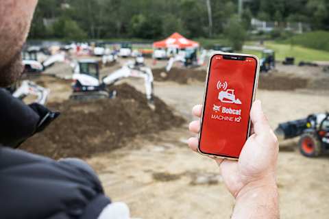 Pendant les Demo Days de Bobcat, un homme tient un téléphone qui affiche l’écran de démarrage de Machine IQ, avec des machines exposées en arrière-plan.