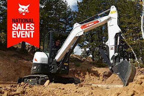 A Bobcat E35 Compact Excavator With a Bucket Digs a Hole in the Dirt Near Trees; the National Sales Event Banner Is on the Top-Left Corner
