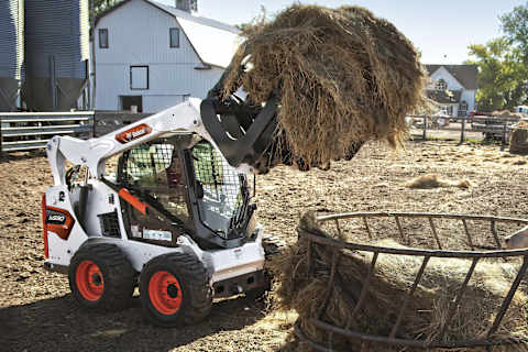 Operatore che usa una pala compatta gommata Bobcat S590 con pinza per radici per scaricare un grande cumulo di fieno in una mangiatoia in una fattoria.
