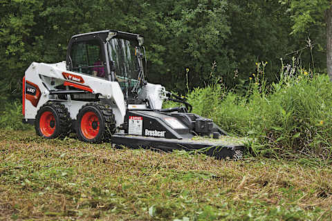 Operatrice che usa la pala compatta gommata Bobcat S510 con accessorio per silvicoltura Brushcat per togliere le sterpaglie in una zona alberata.