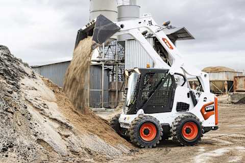 Opérateur aux commandes d’une chargeuse compacte sur pneus S630 de Bobcat pour décharger un godet de sable sur un tas plus important par temps nuageux.