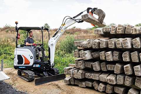 Un uomo usa un miniescavatore elettrico Bobcat E19e compatto per sollevare un una traversina in calcestruzzo da una pila in un cantiere di costruzione esterno. Sullo sfondo della scena c'è un terreno erboso e il cielo parzialmente nuvoloso.