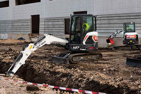 Two Bobcat E27 compact excavators working on a muddy construction site next to a modern building. The excavator in the foreground is digging a trench, while the one in the background is lifting a large rock with a clamp attachment. Both operators are wearing high-visibility safety vests, and the area is marked off with caution tape.