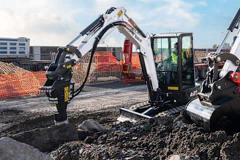 A Bobcat E27 compact excavator using a Bobcat B680 hydraulic breaker attachment to break rocks at a road construction site. The operator is inside the enclosed cab, and the area is surrounded by safety orange fencing, trucks, and construction materials under a bright, partly cloudy sky.