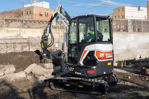 A Bobcat E27 compact excavator equipped with a hydraulic breaker attachment demolishing concrete blocks on an urban construction site. The operator is inside the cab, and the background features a temporary site office, a concrete wall, and residential buildings under a clear blue sky.