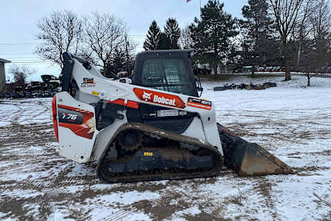 The recovered stolen Bobcatâ T76 compact track loader with snowblower attachment parked with bare winter trees in the background and with an American flag on a flag pole. 