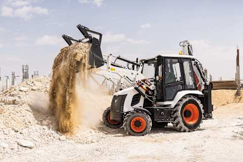 Bobcat B730M backhoe loader in action on a dusty, sunlit construction site in Dubai, captured mid-motion as it dumps a full bucket of dirt against a backdrop of arid terrain and development.