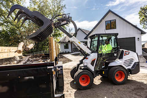 A Bobcat L35 Small Articulated Loader Uses a Root Grapple Attachment to Dump Logs Into a Trailer on a Residential Job Site