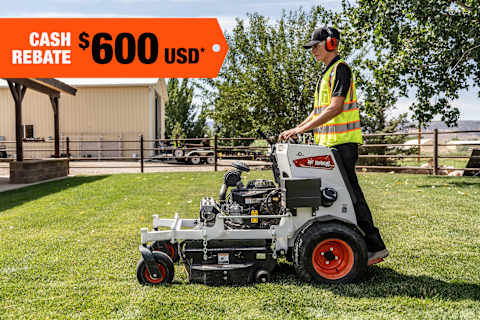 An Operator Mows a Fenced-In Area With a Bobcat Zero-Turn Stand-On Mower; A Cash Rebate Banner Is in the Top-Left Corner