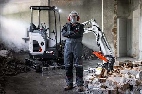 A worker in protective gear stands before a Bobcat E17z mini excavator inside a partially demolished building with brick debris.