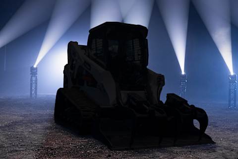 A Bobcat T650 Compact Track Loader Equipped With an Industrial Grapple Is Positioned on a Dirt Surface in a Dramatically Lit Industrial Settin