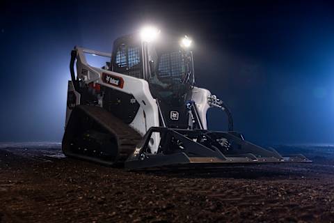 An Operator Grades a Dirt Surface at Night Using a Bobcat T76-2 Compact Track Loader With A Landplane Attachment Under Bright Work Lights Illuminating The Site