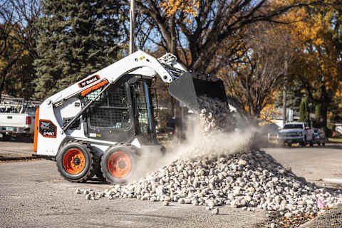 A Bobcat Classic S450 Skid-Steer Loader Uses a Bucket to Dump a Pile of Rocks on a Residential Street During Landscaping Work