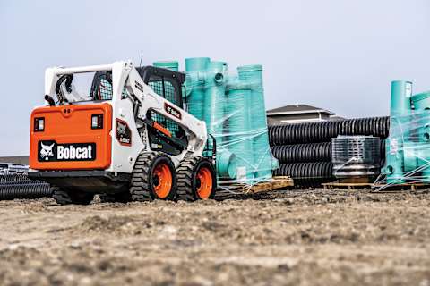A Bobcat Classic S590 Skid Steer Loader with Pallet Fork Attachment Moves Stacked Pipes on Pallet at a Construction Site
