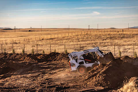 A Bobcat Classic S770 Skid-Steer Loader Uses a Bucket to Move Dirt Across a Jobsite With Open Fields in the Background