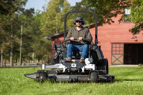 Front angle closeup shot of Bobcat zero-turn riding mower with male operator mowing large property with red barn in the background
