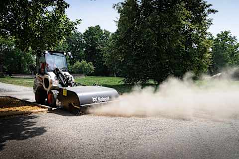 Bobcat small articulated loader with a sweeper attachment kicking up dust on a park path