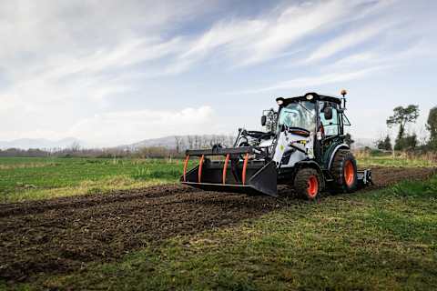 Bobcat compact tractor with FL8 front loader grapple bucket and rear tiller preparing soil in a field