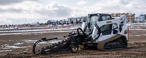 An Operator Digs a Narrow Trench Using a Bobcat T770 Compact Track Loader With a Trencher Attachment on a Snow-Dusted Construction Site Near Residential Buildings