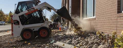 A Bobcat Classic S70 Skid-Steer Loader Uses a Bucket to Move Rocks and Debris Along a Building Foundation During Landscaping Work