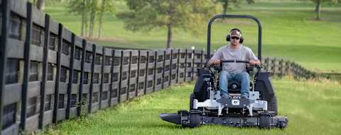 Bobcat ZT6100 zero-turn riding mower with male operator mowing large property with large acreage field and trees in the background