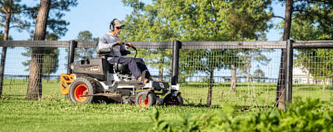 Bobcat ZT3000 zero-turn riding mower in action mowing along fence line with trees and large acreage farm property in the background
