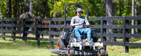 Bobcat ZT6000 zero-turn riding mower in action with male operator mowing large horse ranch property with fence, horses and trees in the background