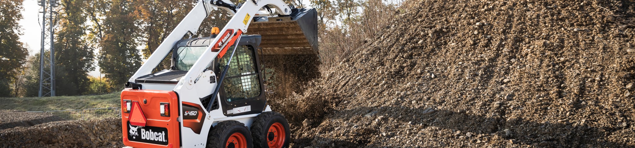 Une chargeuse compacte sur pneus Bobcat vue de l’arrière, ses balanciers étendus inclinés vers une pile, pendant des travaux d’aménagement paysager par une journée ensoleillée.