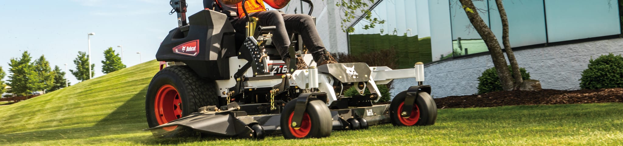 Un operador utilizando una segadora Bobcat de giro cero para cortar el césped junto a un gran edificio en un día soleado.