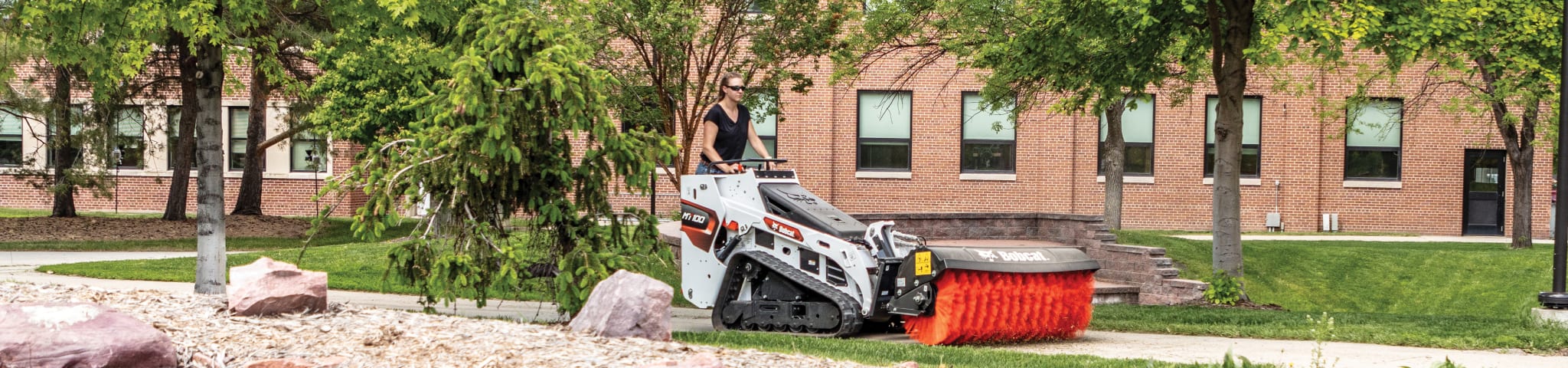 Eine weibliche Bedienerin, die einen Bobcat-Mini-Raupenlader mit dem Schwenkbesen als Anbaugerät verwendet, um einen Weg vor einem großen Gebäude von Unrat zu befreien.