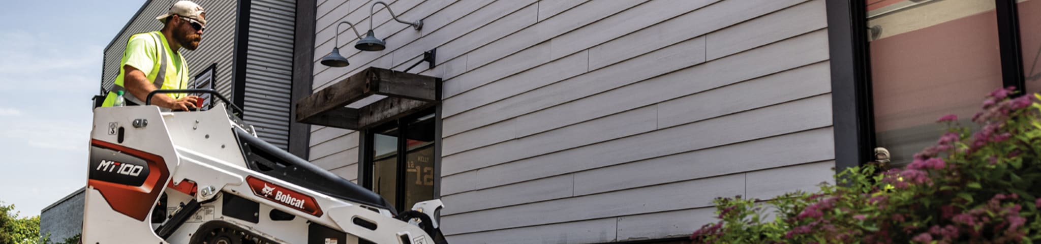 An Operator Uses a Bobcat MT100 Mini Track Loader With a Bucket Near a Building