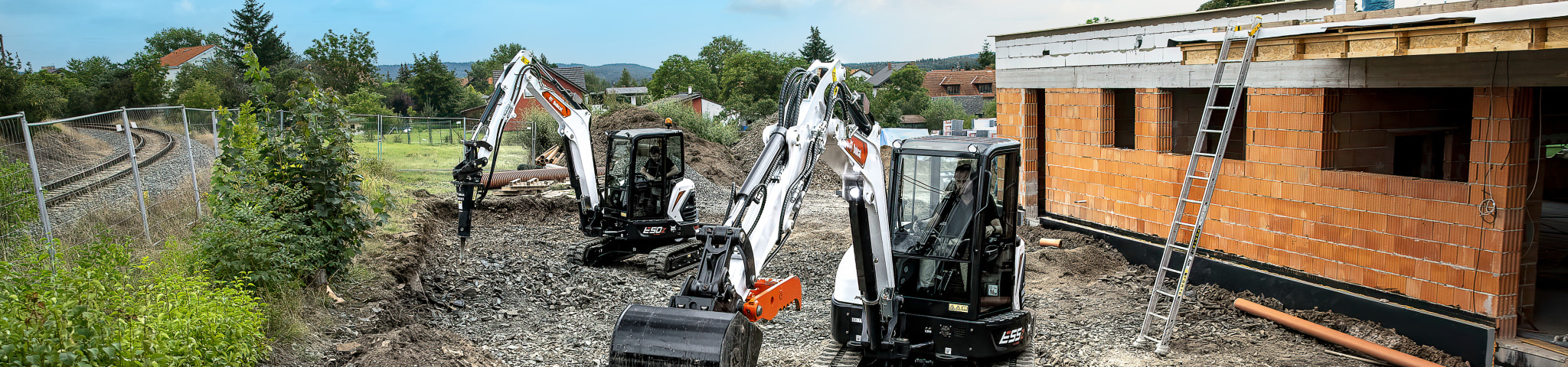 Two male operators in tandem operating two Bobcat mini excavators as they navigate the rubble-strewn terrain of a housing project.