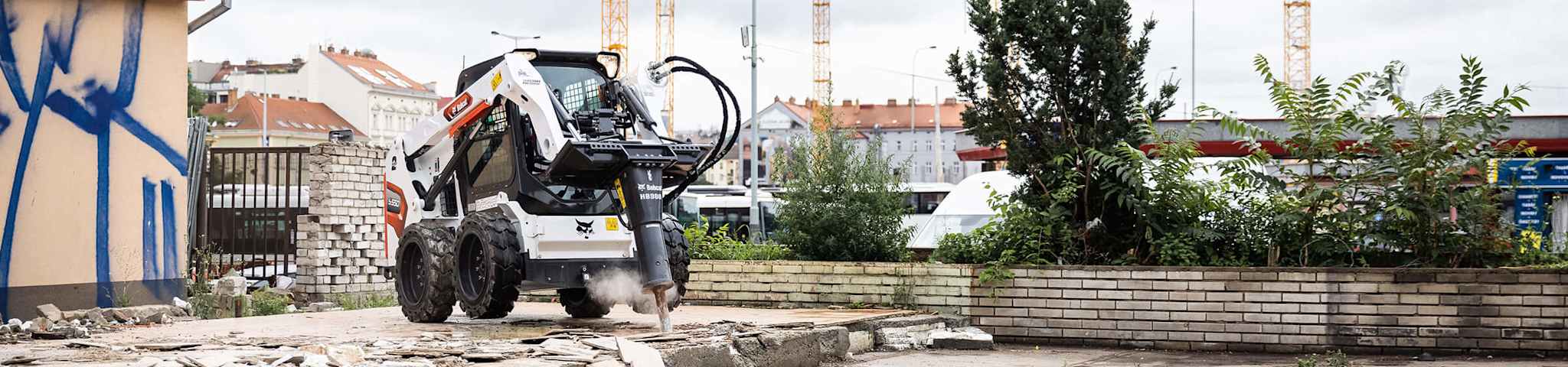 A Bobcat skid-steer with a hydraulic breaker demolishes concrete on an urban jobsite, with cranes and buildings in the background.