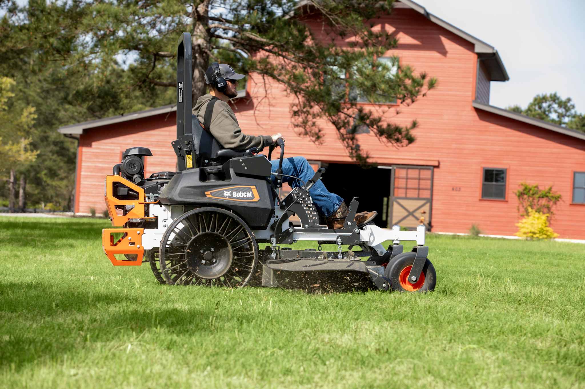 Side profile action shot of male operator mowing with a Bobcat ZT7000 zero-turn riding mower with grass clippings flying out of the discharge chute and large red barn in the background
