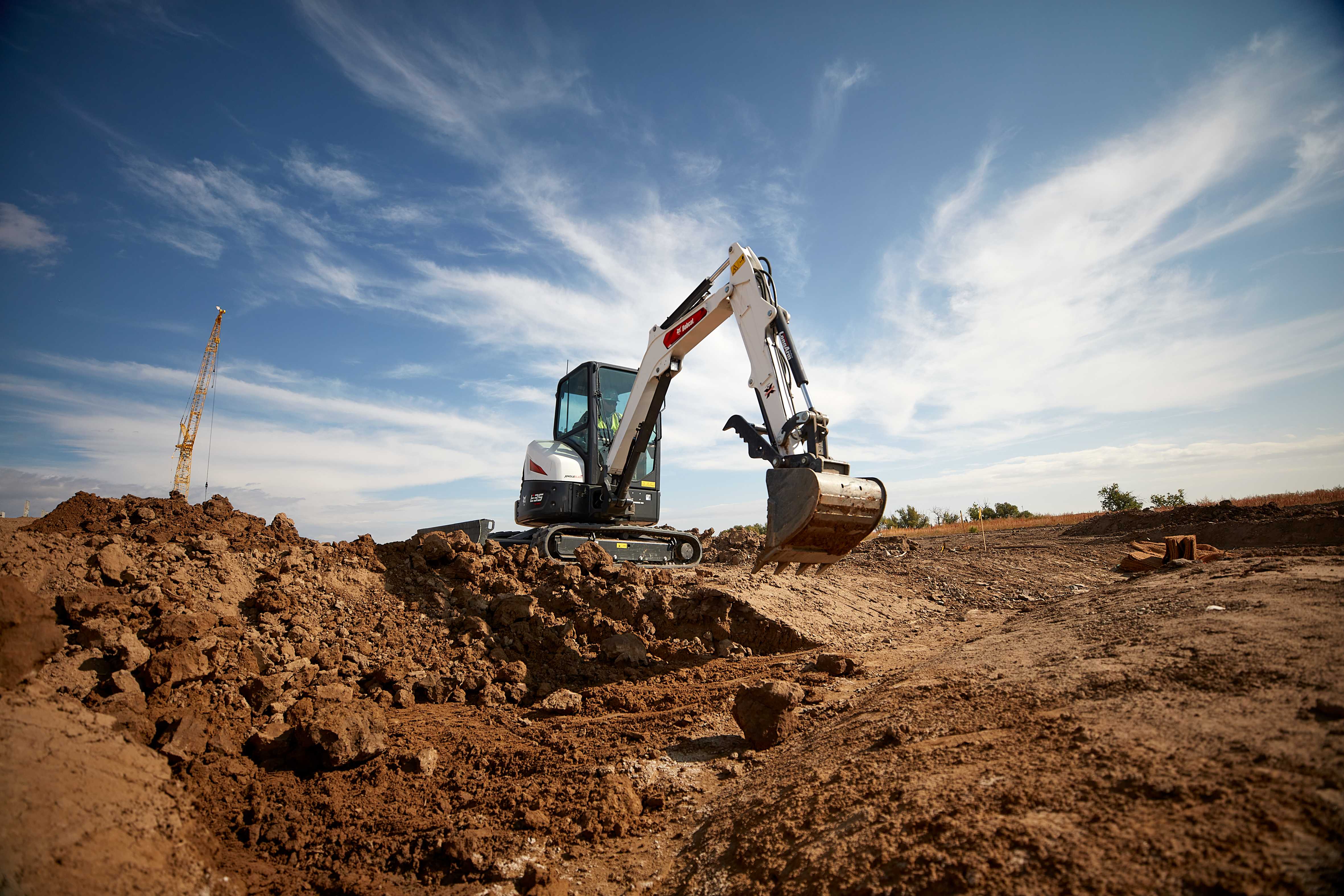 Bobcat E35 Compact Excavator with Bucket Attachment Excavating Soil at a Construction Site With a Crane in the Background