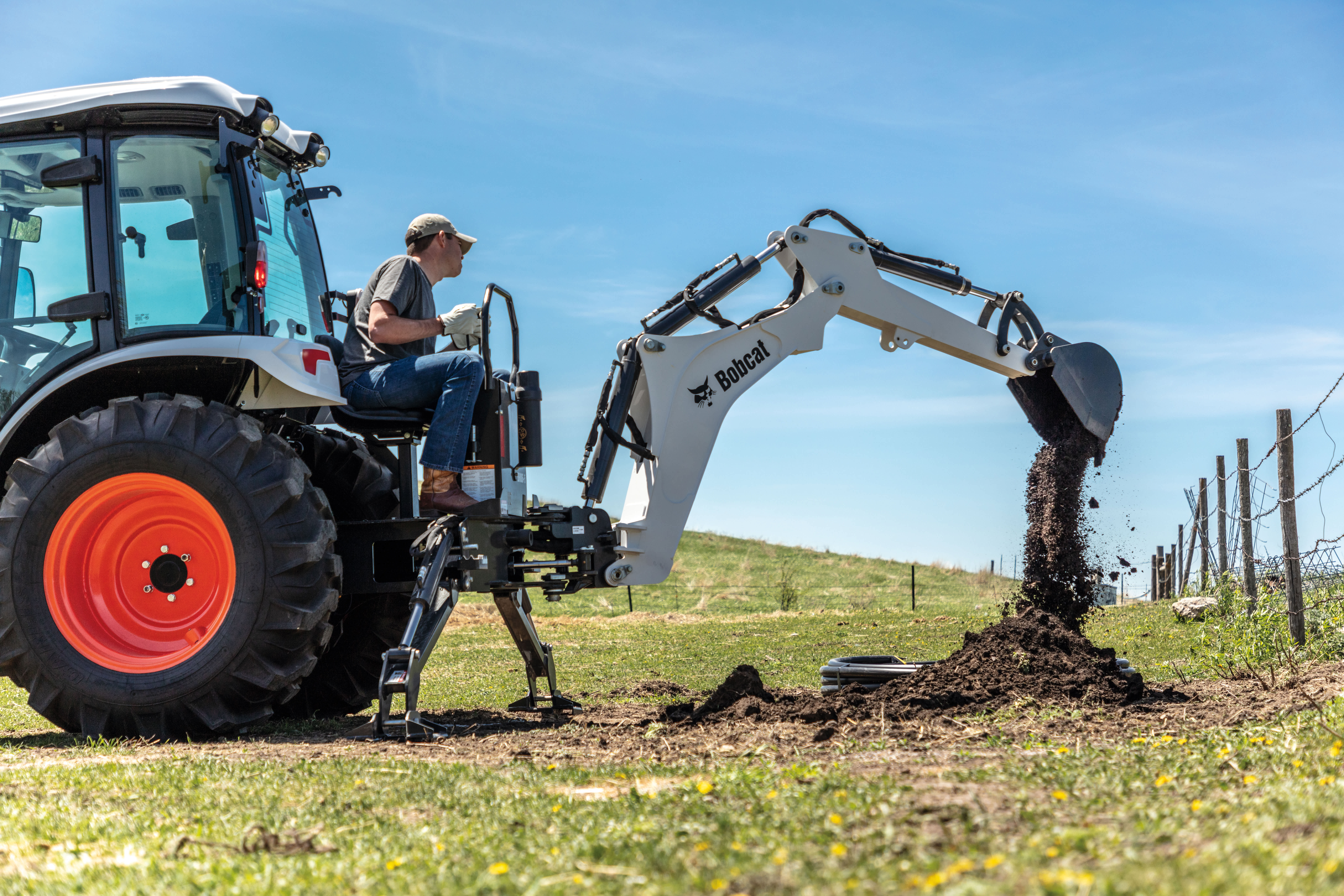An Operator Digs a Hole Near a Fence Line Using a Bobcat Compact Tractor Equipped With a Backhoe Attachment 