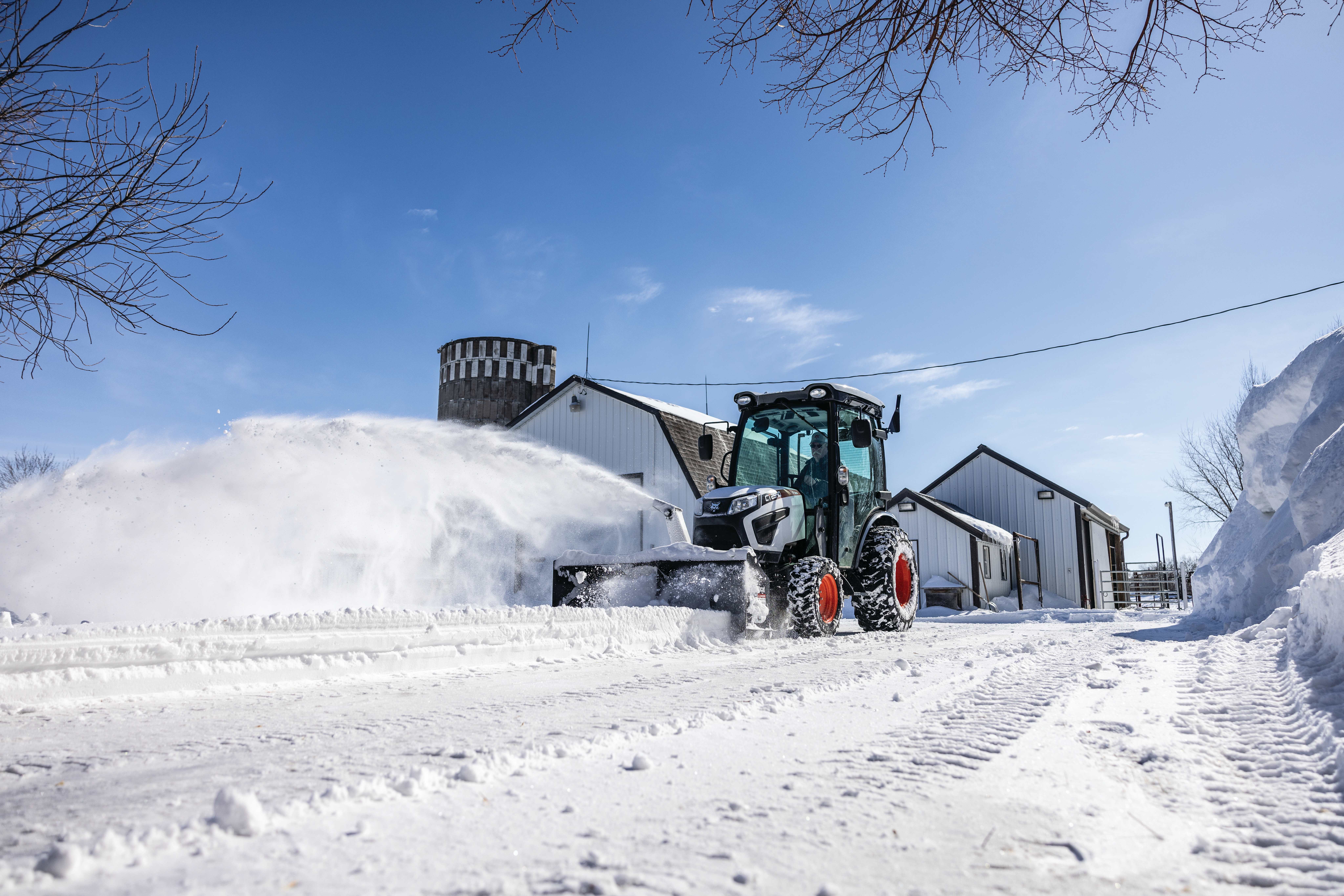 An Operator Clears Snow From a Farm Driveway Using a Bobcat CT2540 Compact Tractor With a Front-Mount Snowblower Attachment Near Agricultural Buildings