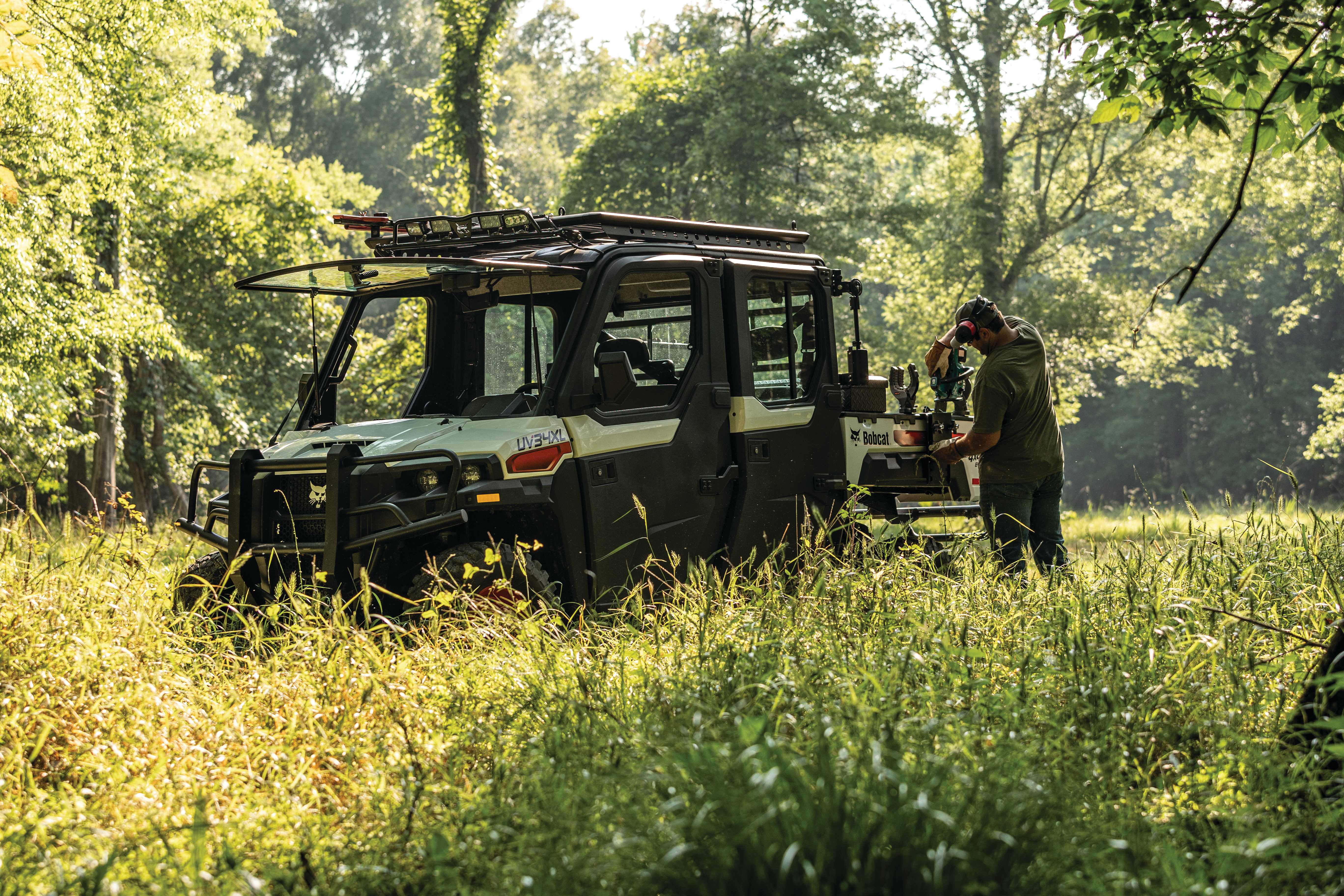 Bobcat UV34XL UTV parked in wooded area with tall grasses and surrounded by trees with operator grabbing chainsaw from the bed of the vehicle